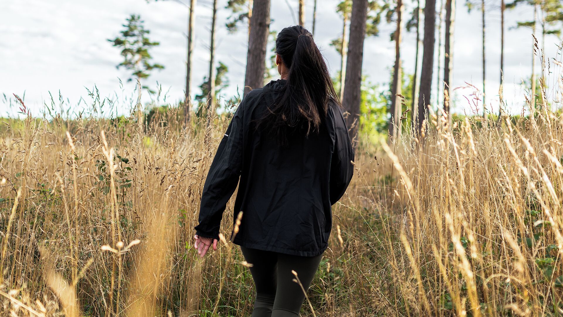 A person walking through a field with tall grasses and trees in the background.