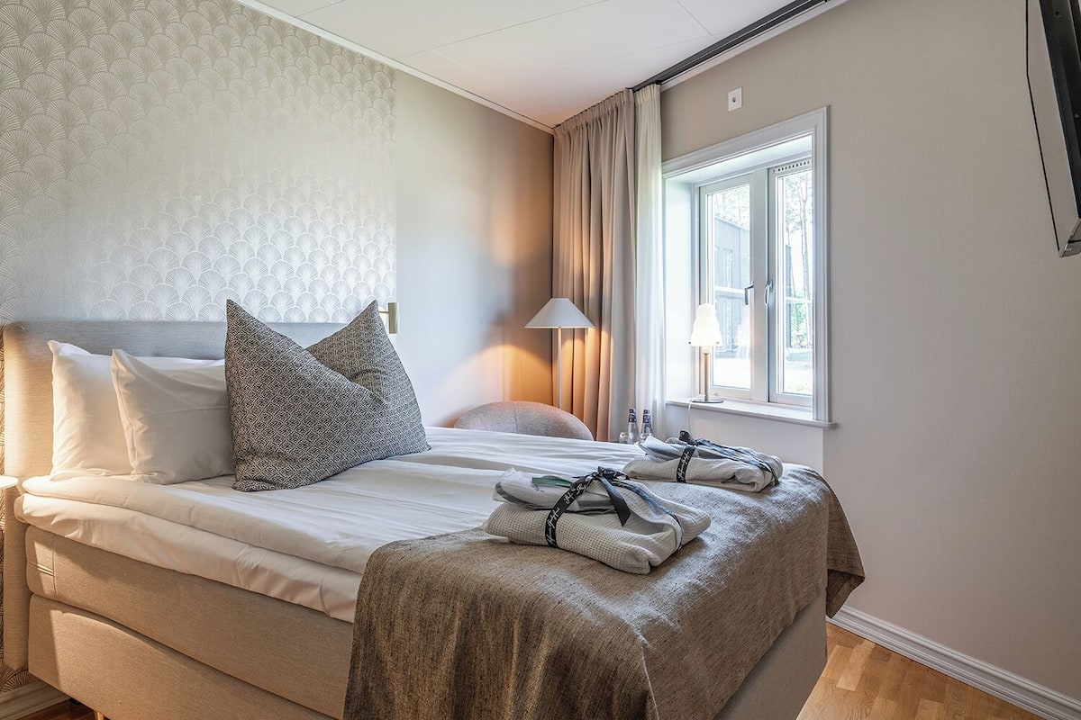 A hotel room with a bed, pillows, a bedside lamp, and a window with curtains.