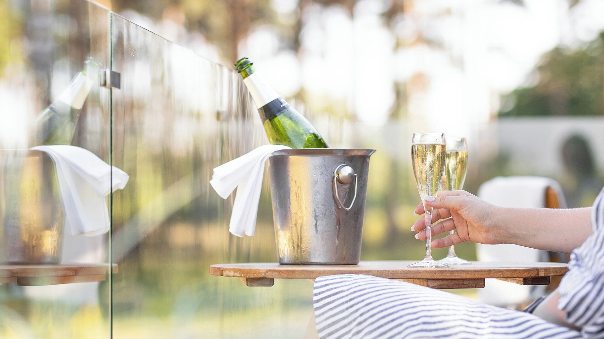 A person holding two champagne glasses, a bucket with a bottle, and a white cloth on a wooden table outdoors.