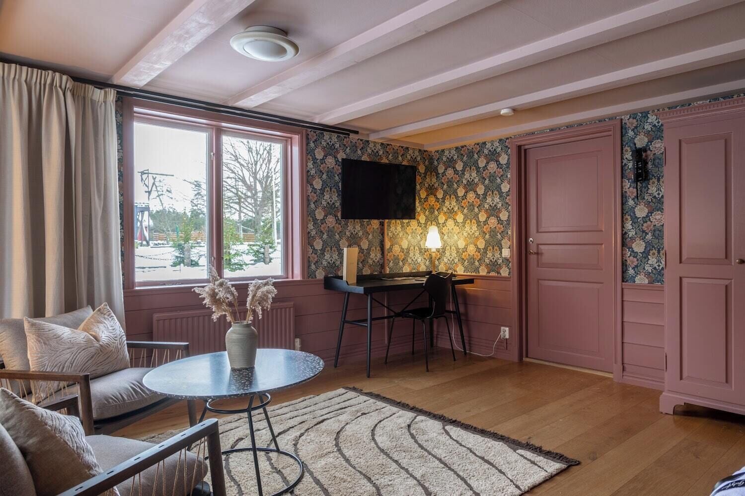 Una habitación con una mesa redonda, una silla, un televisor montado en la pared y una puerta con papel tapiz floral.