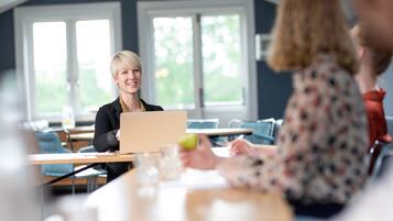 A conference room with a laptop on a table, a person holding a green apple, and another person in the background.
