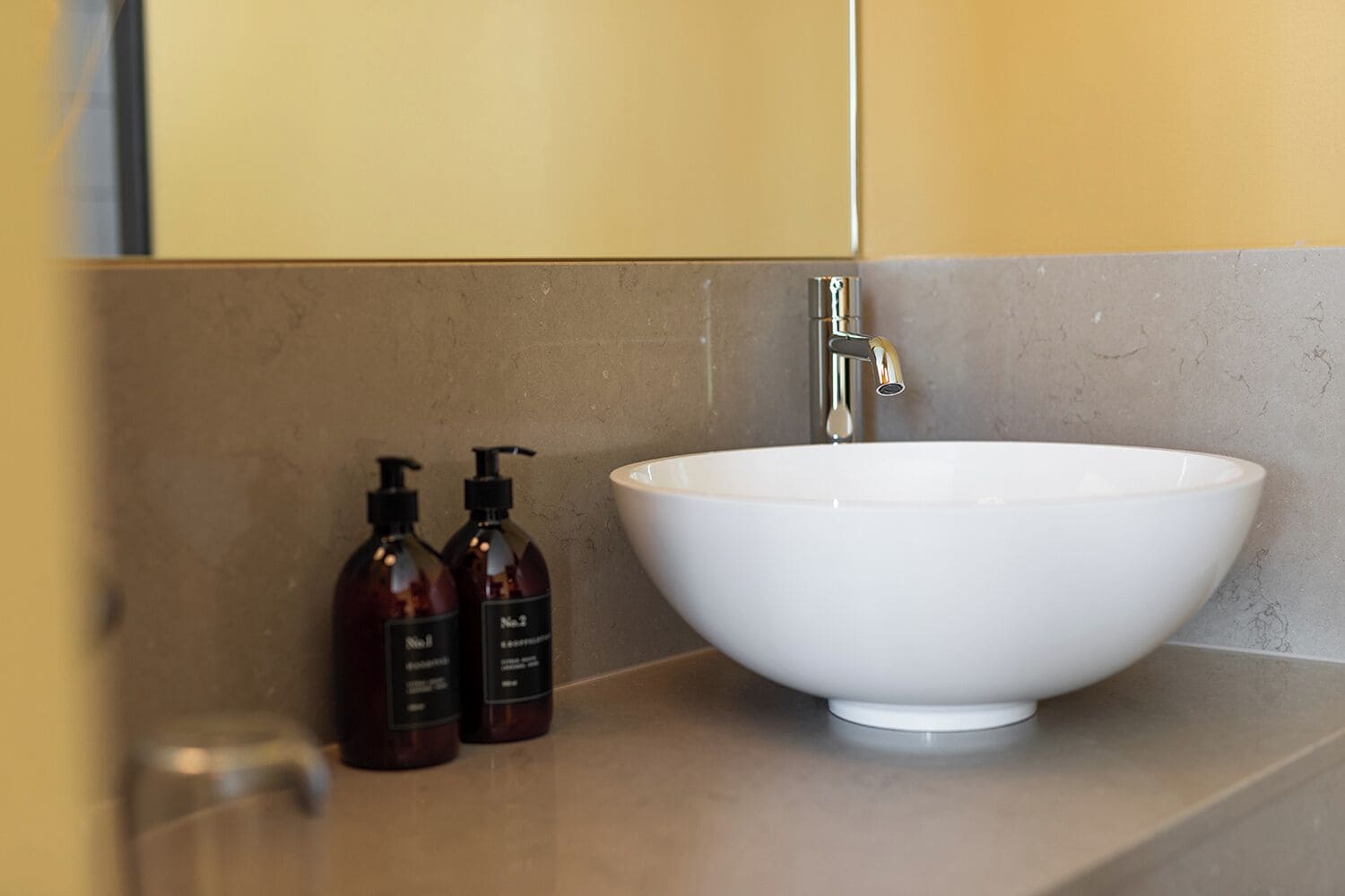 A bathroom with a white vessel sink, a faucet, and two dark brown bottles of soap on a beige countertop.