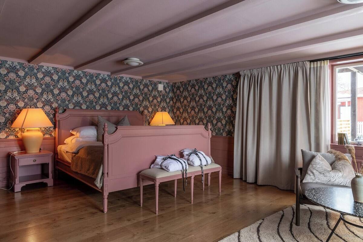 A bedroom with a wooden bed, bedside lamps, a bench, a chair, and a window with curtains.