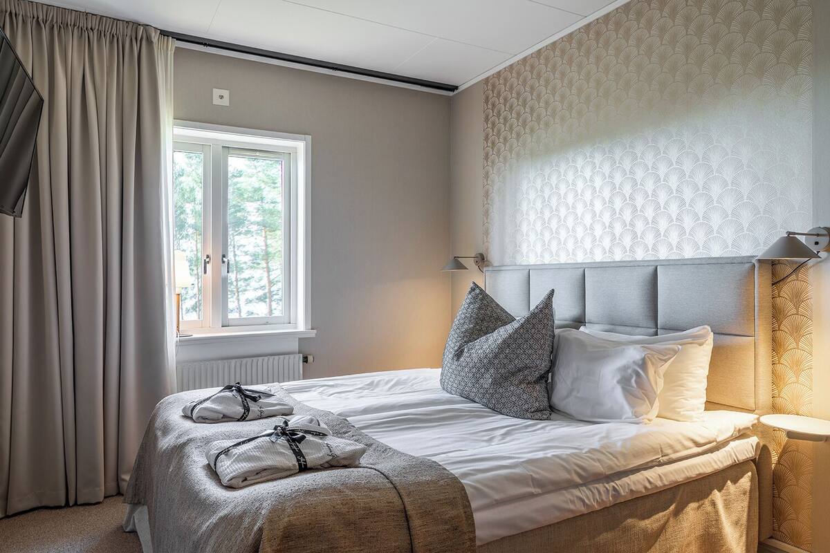 A neatly made bed with a grey headboard, a window with curtains, and a patterned wall.