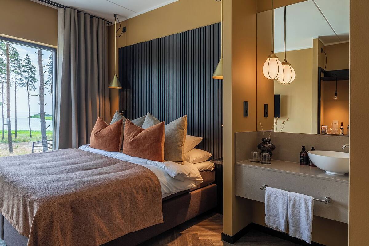 A modern bedroom with a large bed, a bedside table, a mirror, and a view of the outdoors.