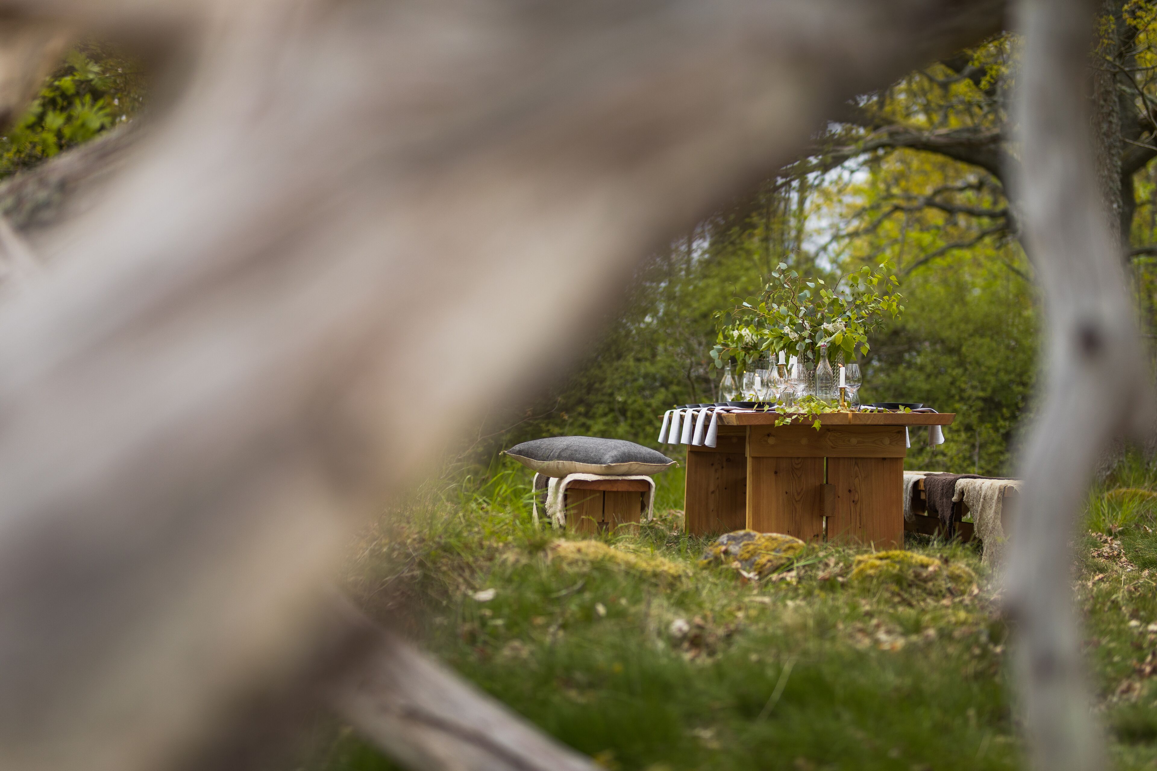 A rustic wooden table with a vase of flowers and a candle, set in a natural outdoor setting with grass and trees.
