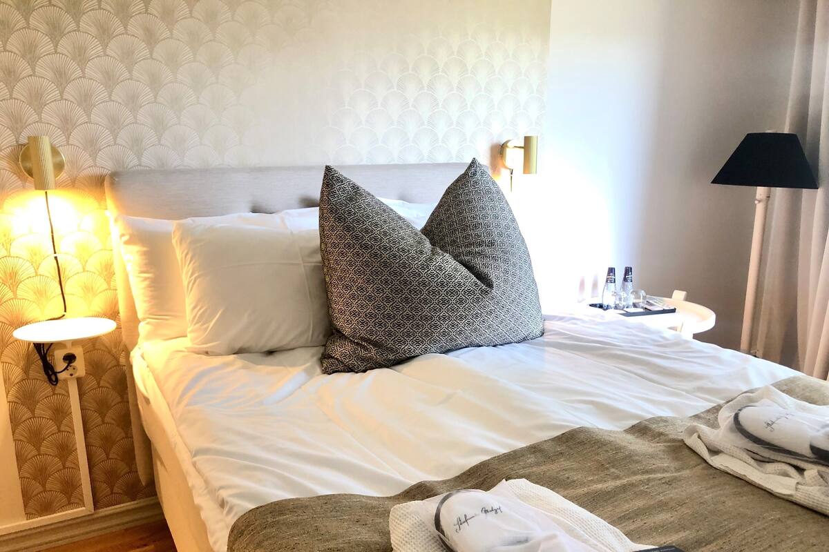 A hotel room with a bed, pillows, a bedside table with a lamp, a nightstand, and a window with curtains.