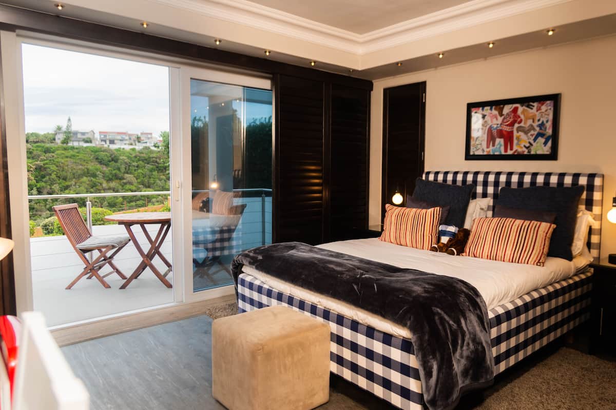 Un dormitorio con una cama grande, ropa de cama a cuadros, un balcón con mesa y sillas, y vistas a la ciudad.