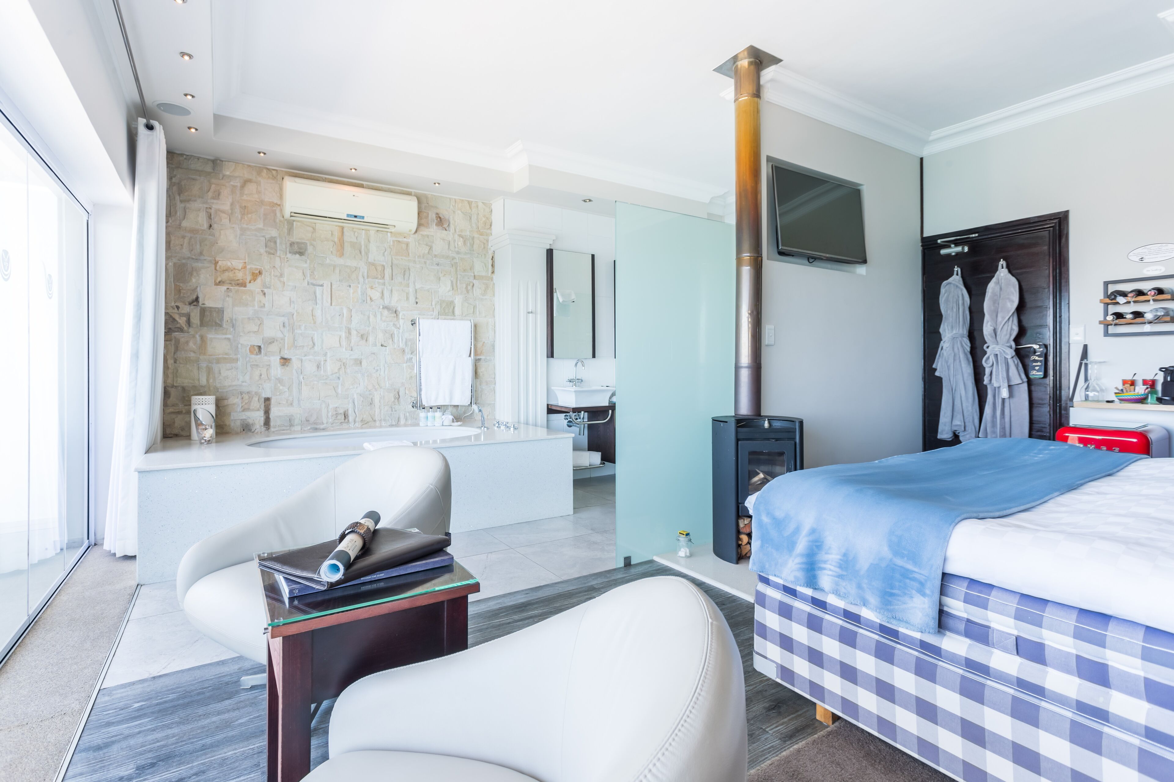 A modern hotel room with a stone wall, a large bed, a fireplace, a TV, and a bathroom with a bathtub and a mirror.