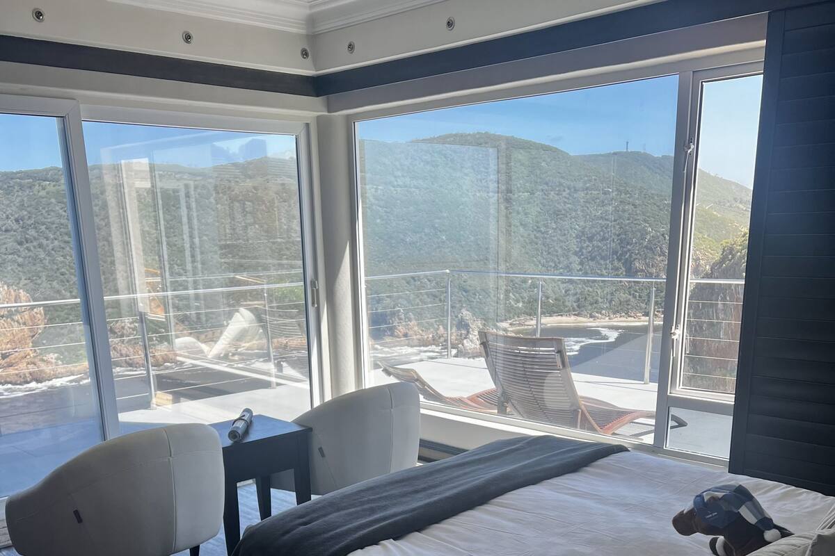 Un dormitorio moderno con una cama grande, un escritorio y dos sillas. Cuenta con un amplio ventanal con vistas a un paisaje montañoso.