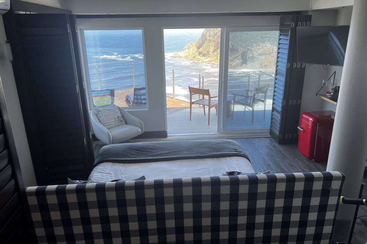 Un dormitorio con cama, sofá a cuadros, refrigerador rojo y puerta corrediza de vidrio que da a un balcón con vista al océano y una terraza de madera.