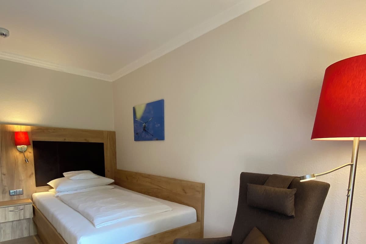 Une chambre moderne avec un lit en bois, un fauteuil gris, une petite table, une lampe de sol rouge et une horloge murale.