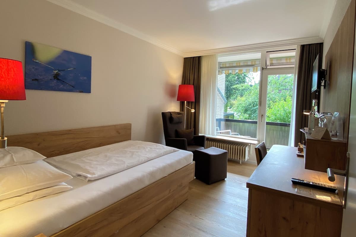 Une chambre d’hôtel moderne avec un lit en bois, un bureau et une chaise. Une fenêtre offre une vue sur la verdure environnante.