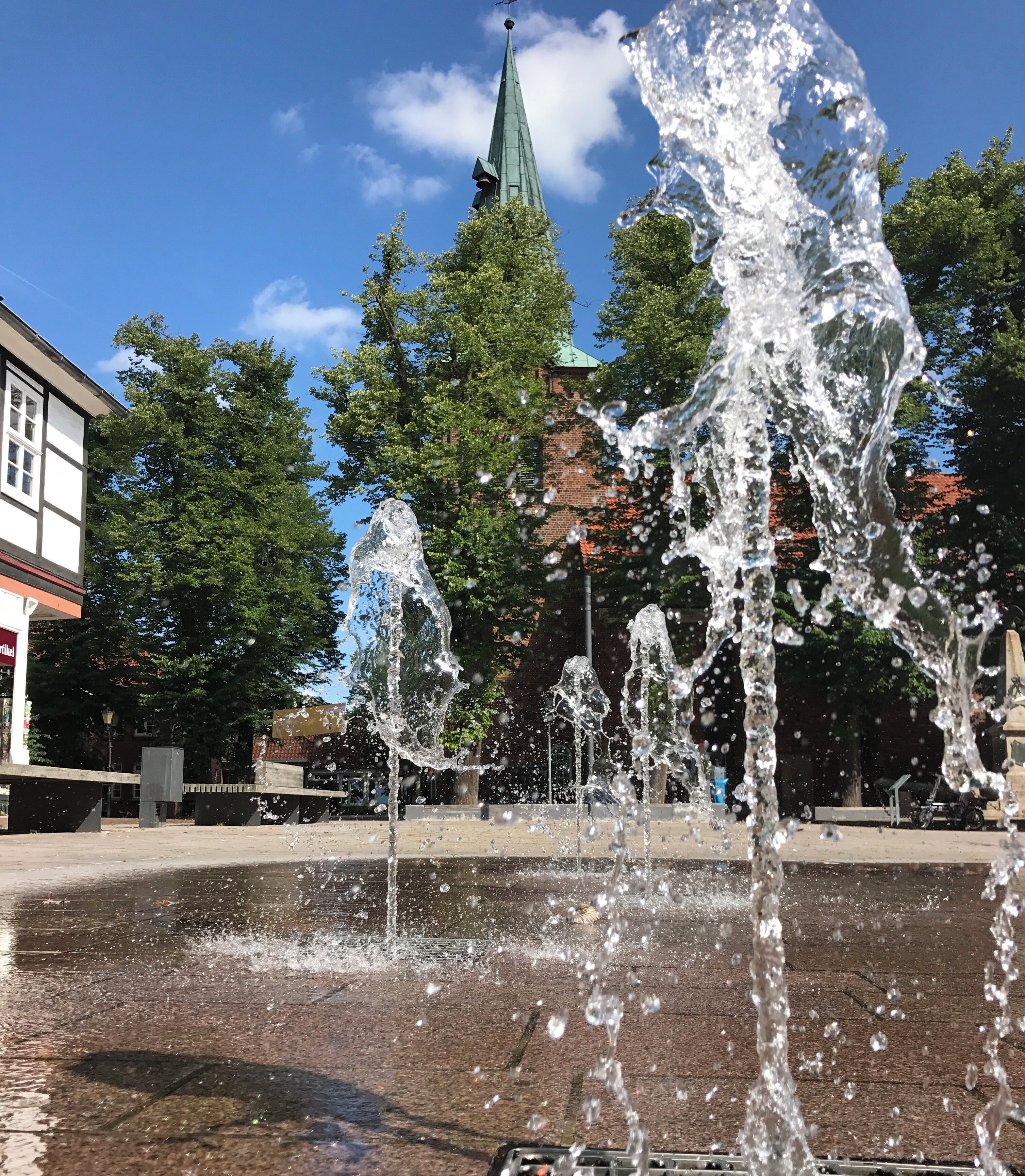 A public fountain with water jets, surrounded by trees and a building with a green roof.
