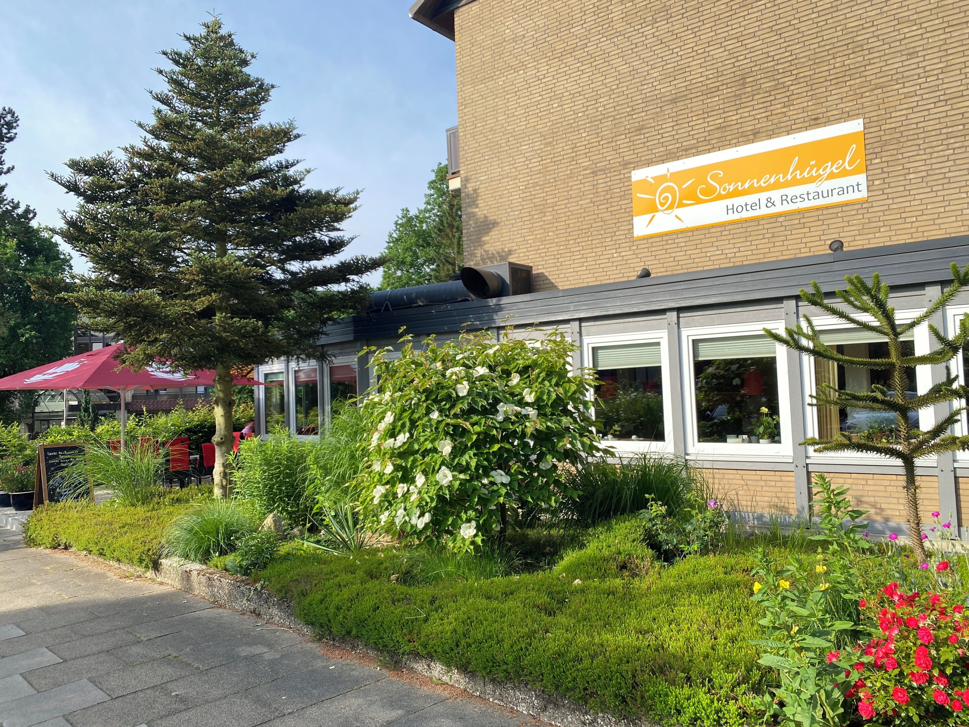 A hotel with a sign reading "Sonnenhügel Hotel & Restaurant" and a garden with various plants and flowers.