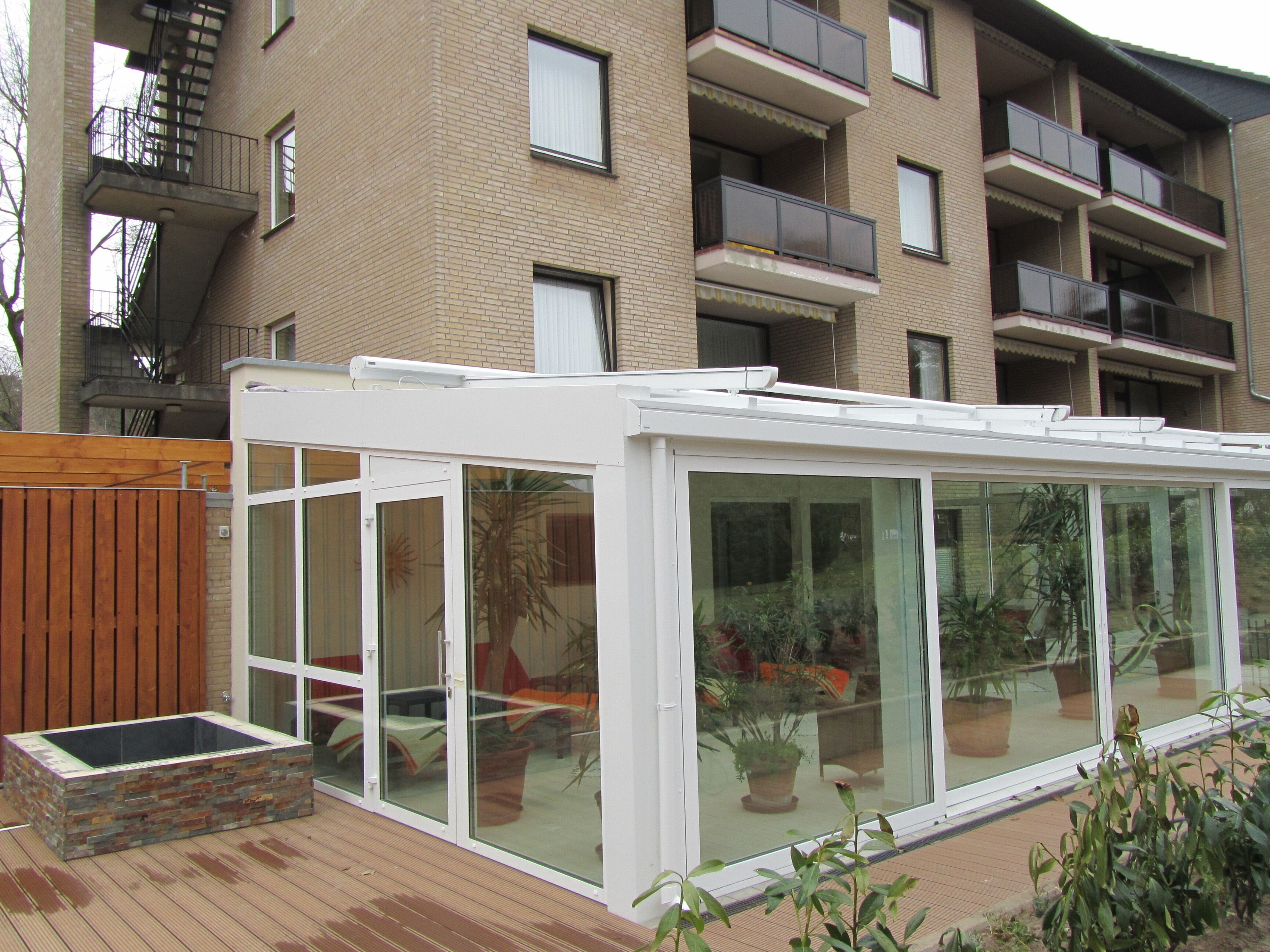 A glass-enclosed patio with potted plants, a wooden deck, and a brick building in the background.