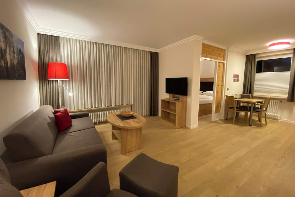Une chambre d’hôtel moderne comprenant un canapé, une petite table basse, un téléviseur, un coin repas avec une table et des chaises, et une grande fenêtre avec des rideaux.