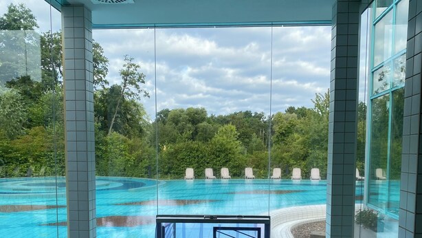 Una piscina interior moderna con paredes de cristal, vistas despejadas del entorno con vegetación y un suelo de baldosas que conduce a la piscina.