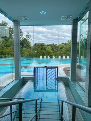 Une piscine intérieure moderne aux murs de verre, offrant une vue dégagée sur la végétation environnante, et dont le sol carrelé mène au bassin.