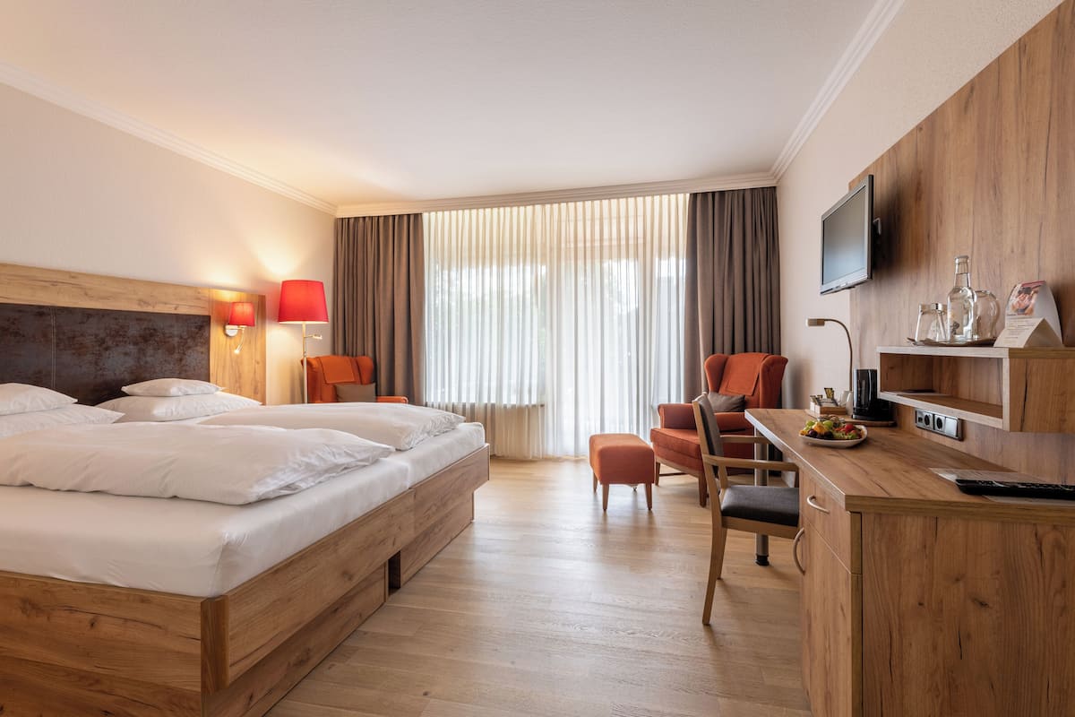 Une chambre d’hôtel avec un lit en bois, un bureau avec une télévision et une grande fenêtre avec des rideaux.