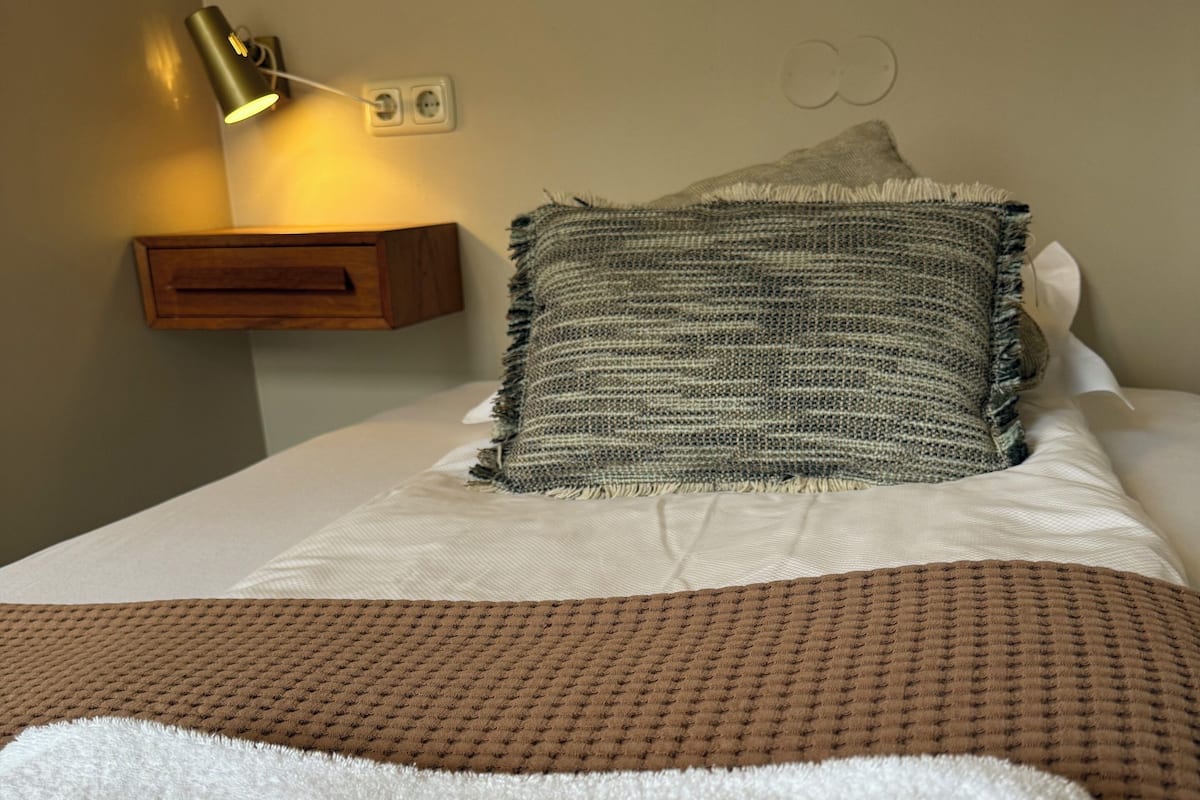 Cama bem arrumada com um cobertor riscado em branco e castanho, um travesseiro e uma luminária de cabeceira.