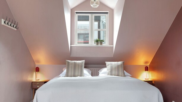 A bedroom with a sloped ceiling, a window with a view, a bed with pillows, bedside lamps, and a wall-mounted light fixture. - Grettir Guesthouse (Reykjavik)