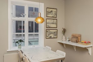 Uma área de refeições com mesa, cadeiras, luminária pendente, janela e prateleiras fixadas na parede com objetos.