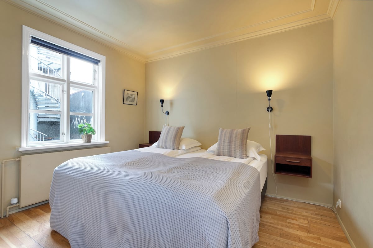 Quarto com cama grande, duas mesas de cabeceira, janela com vista e iluminação embutida na parede.