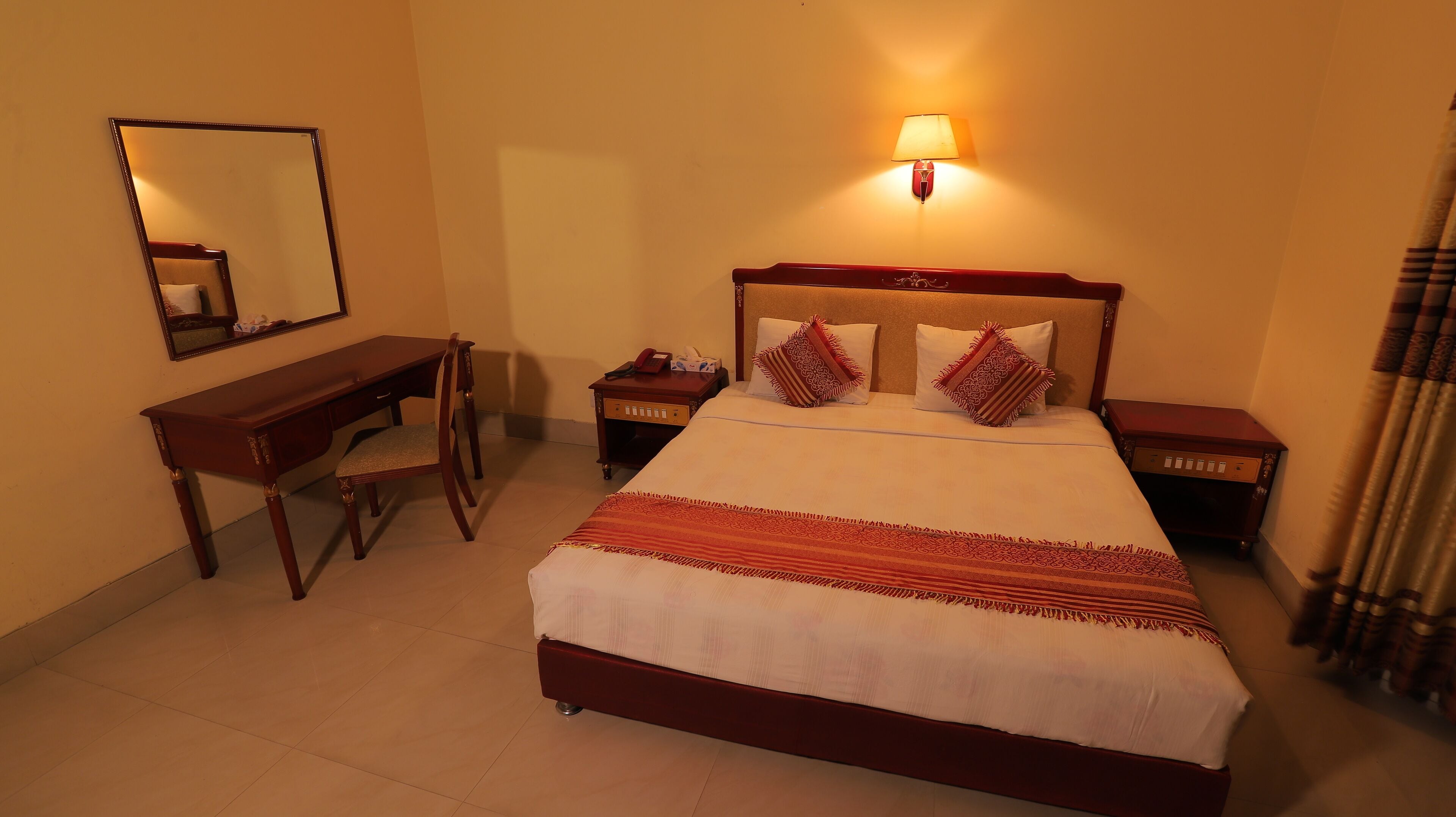 Habitación de hotel con cama, escritorio, silla, mesita de noche y lámpara de pared.