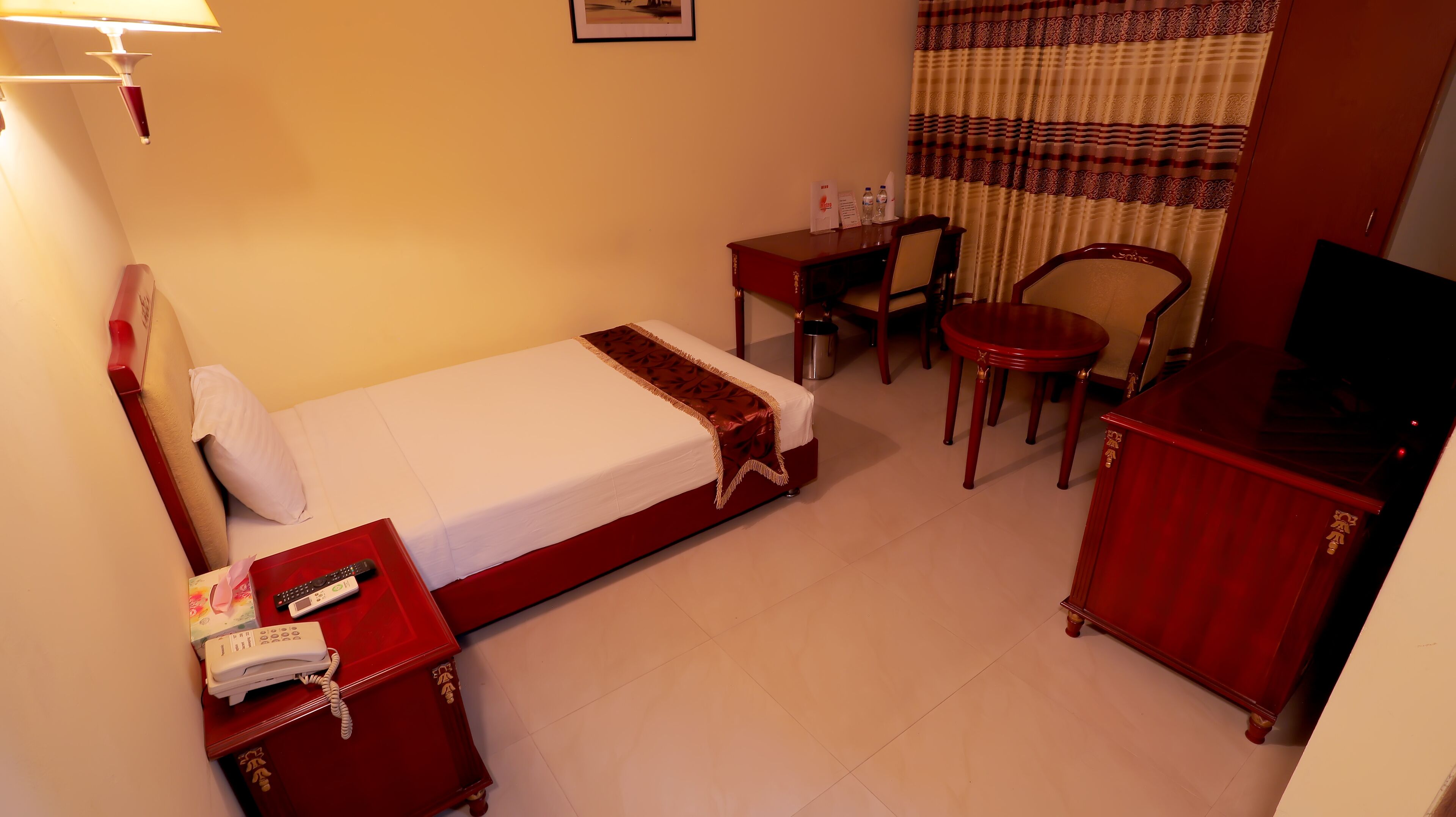 A hotel room with a bed, a desk, a chair, a TV, and a small table with a lamp.