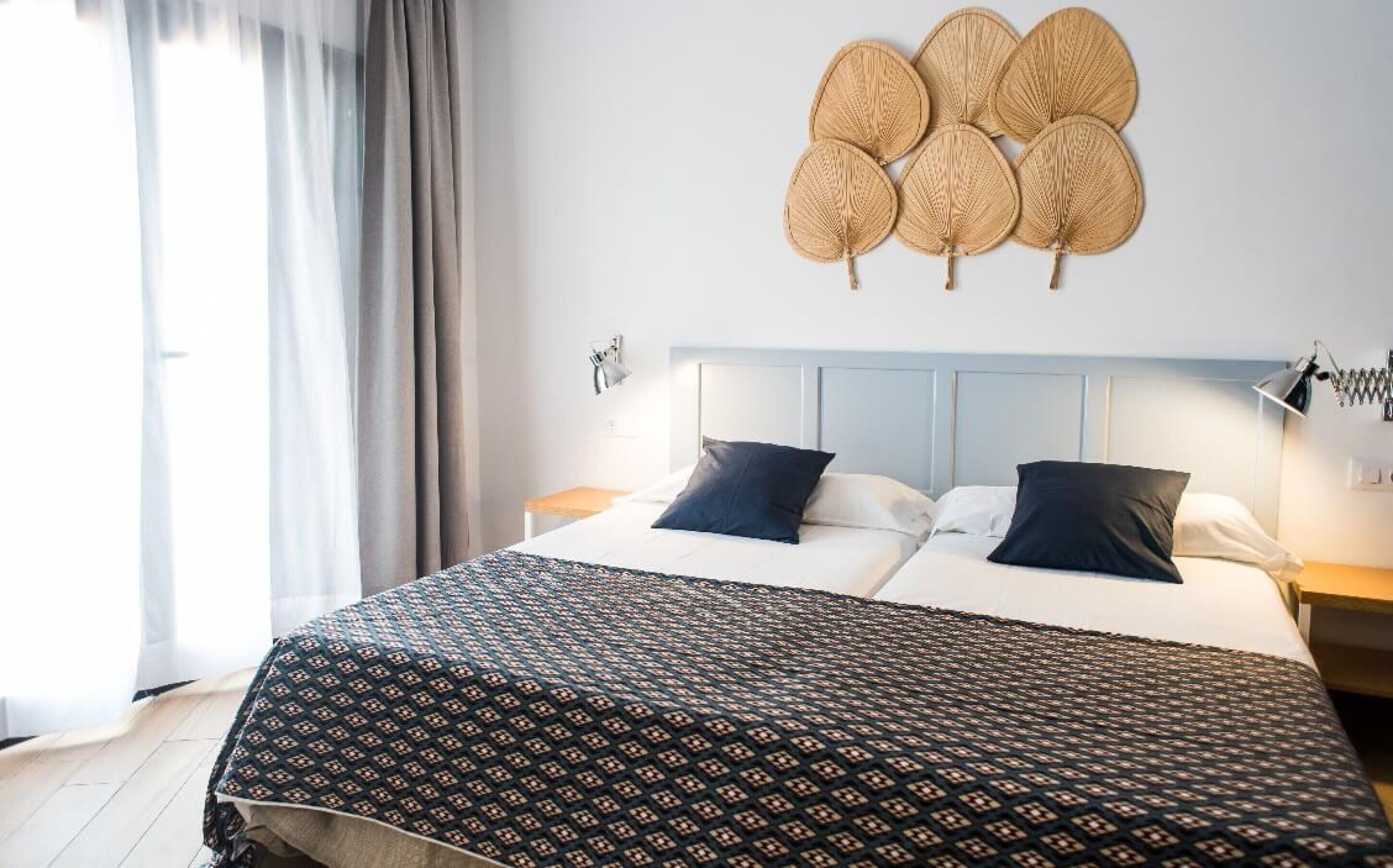 Un lit double avec une couvre-lit à motifs, deux oreillers, une tête de lit, des tables de chevet, une lampe fixée au mur et une fenêtre avec des rideaux.