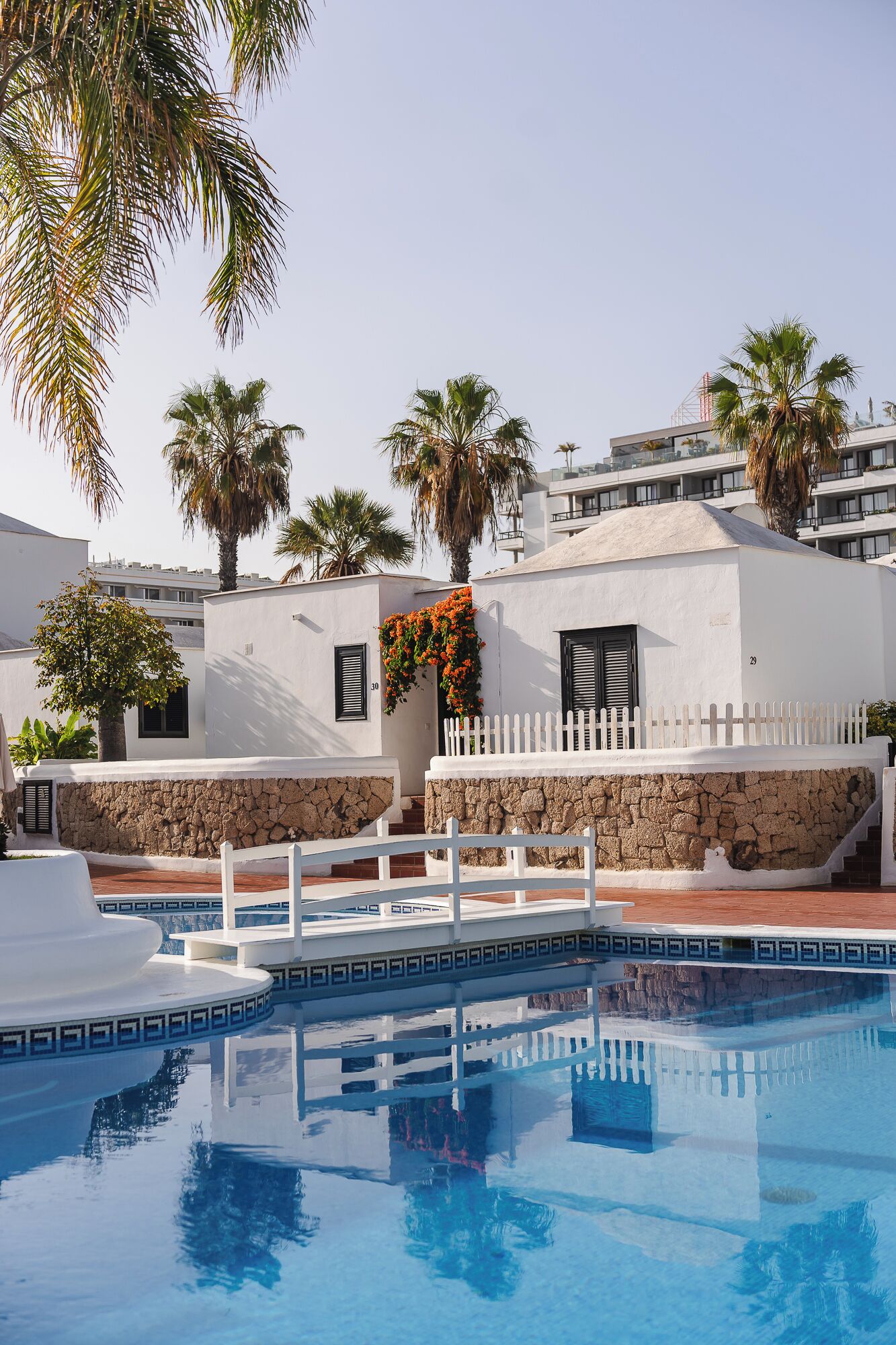 A pool area with a white fence, palm trees, and a building with a stone wall and a white fence.