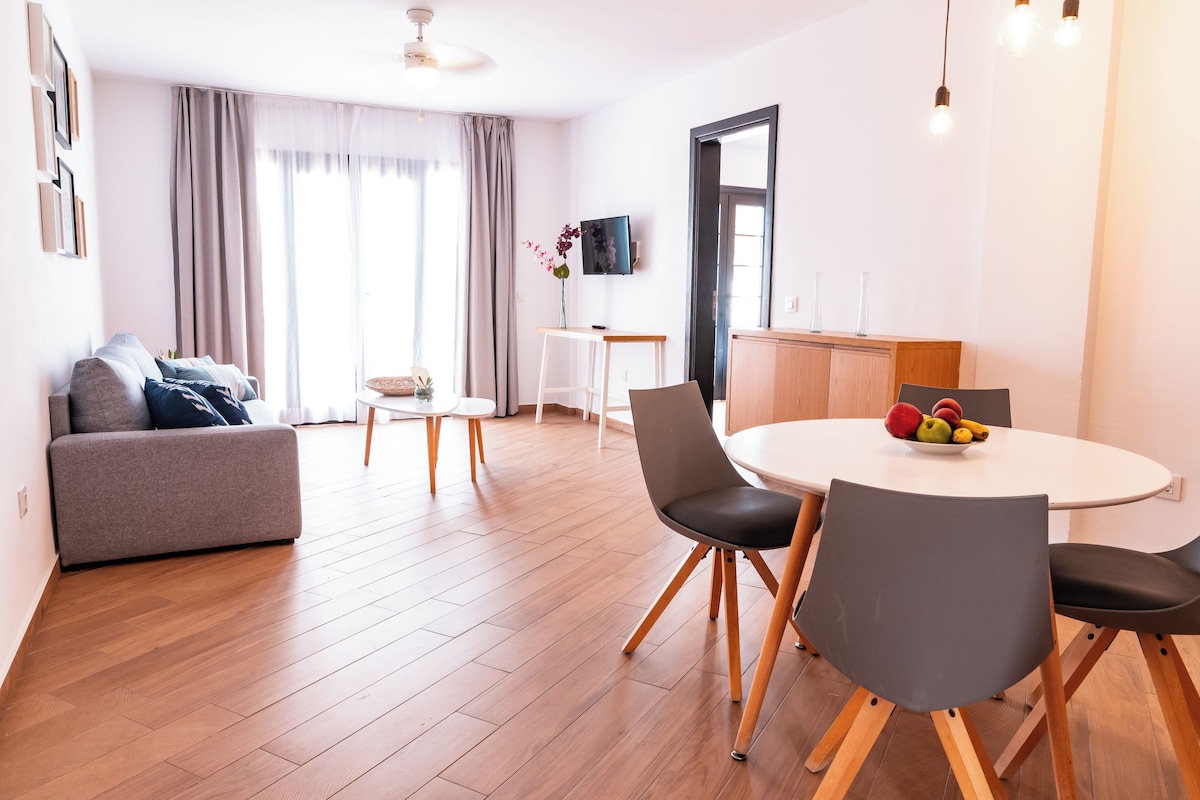 A modern living room with a grey sofa, a dining table with chairs, and a TV mounted on the wall.