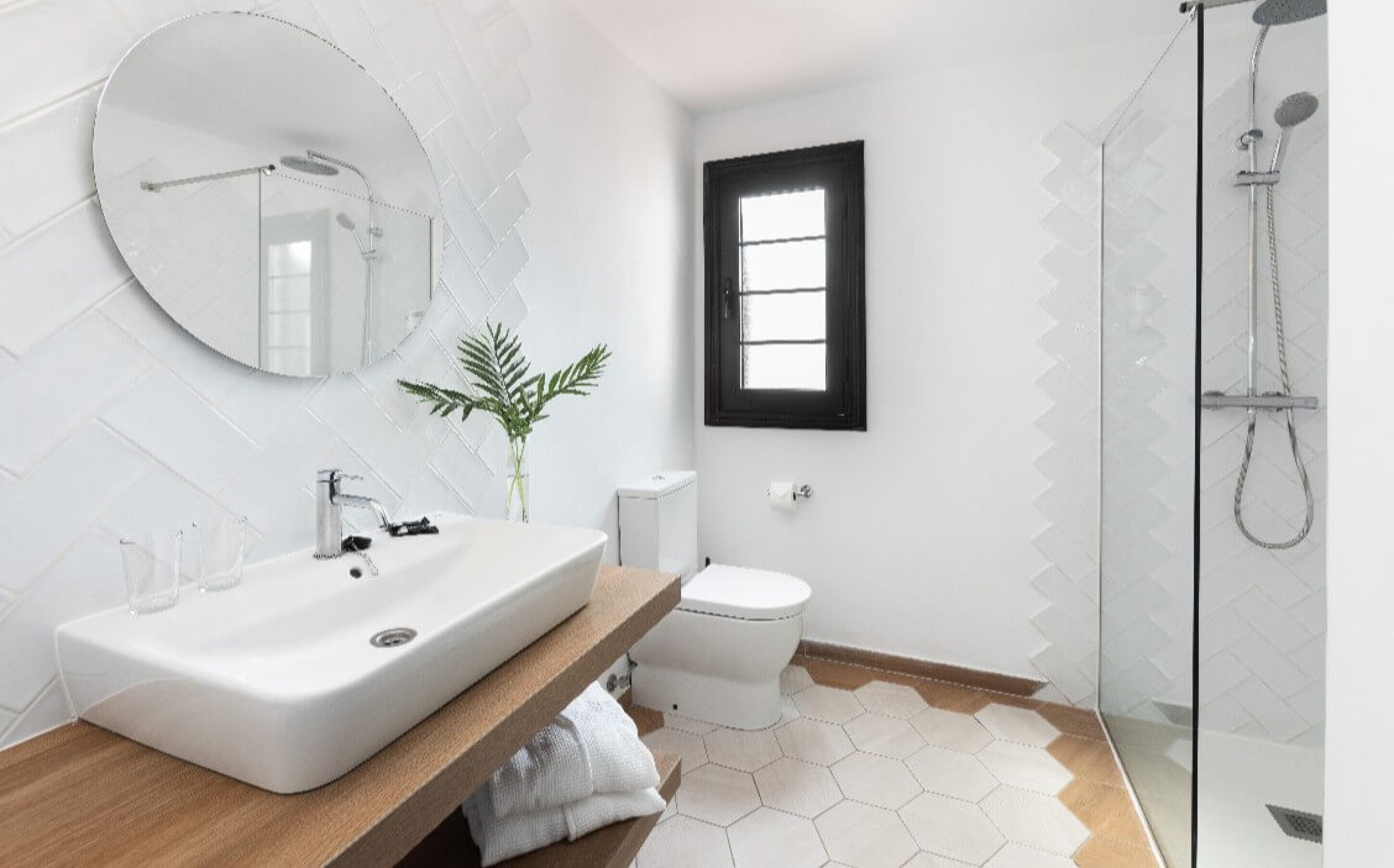 A modern bathroom with a large oval mirror, a white sink, a toilet, and a shower.
