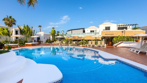 Una piscina con sillas de descanso y sombrillas, rodeada de palmeras y un edificio de recepción.