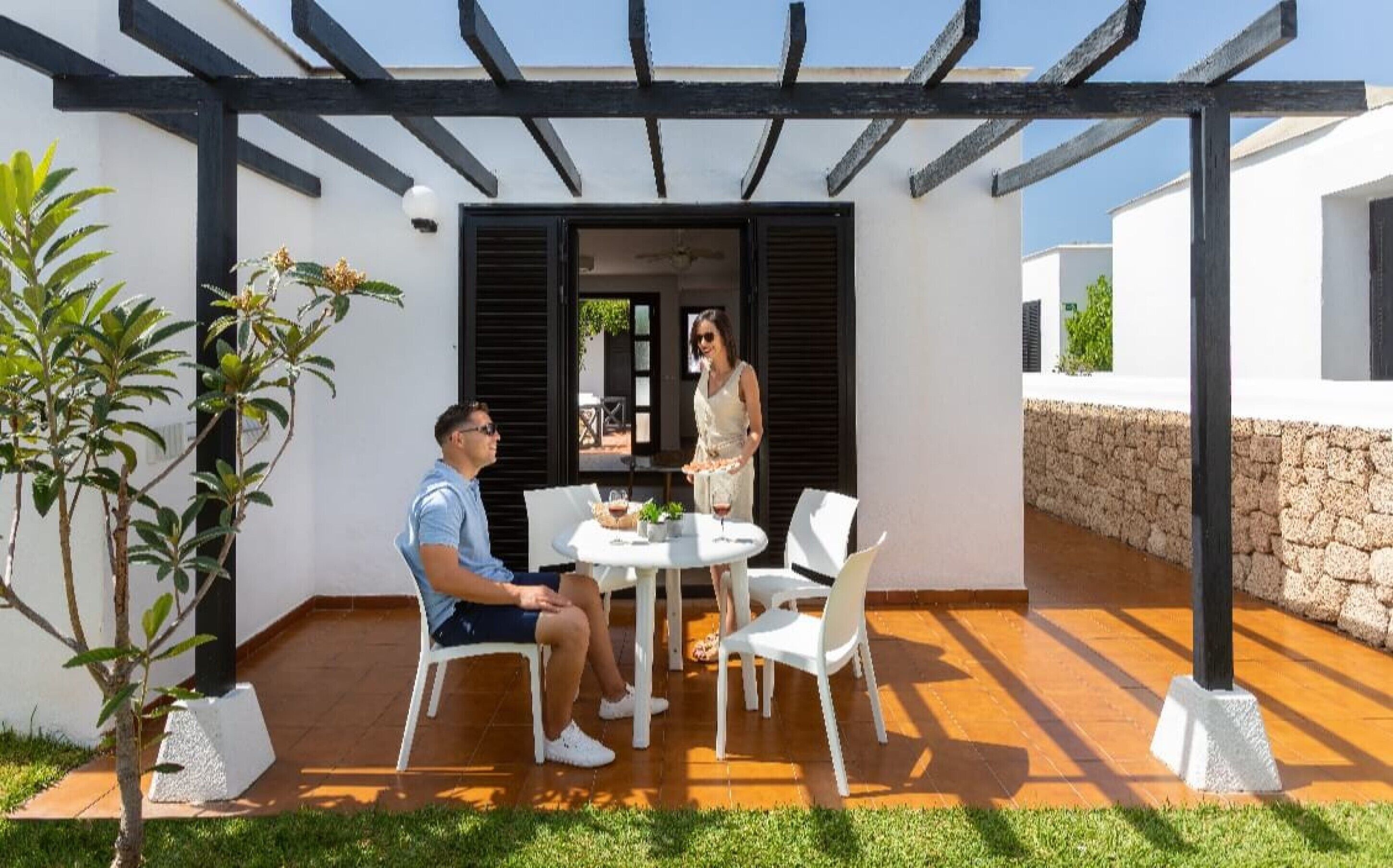 A patio with a pergola, white chairs, and a table.