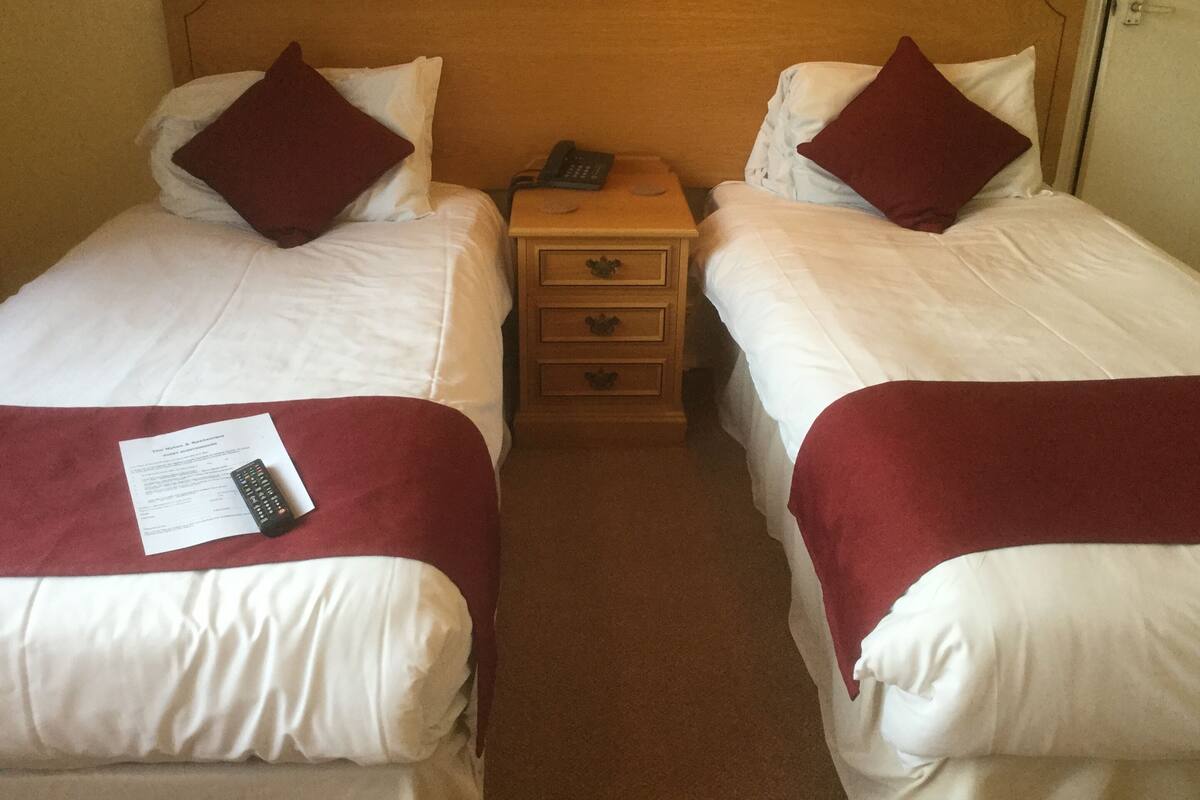 Habitación de hotel con dos camas individuales, cada una con una manta roja, ropa de cama blanca y una mesita de noche de madera entre ellas.