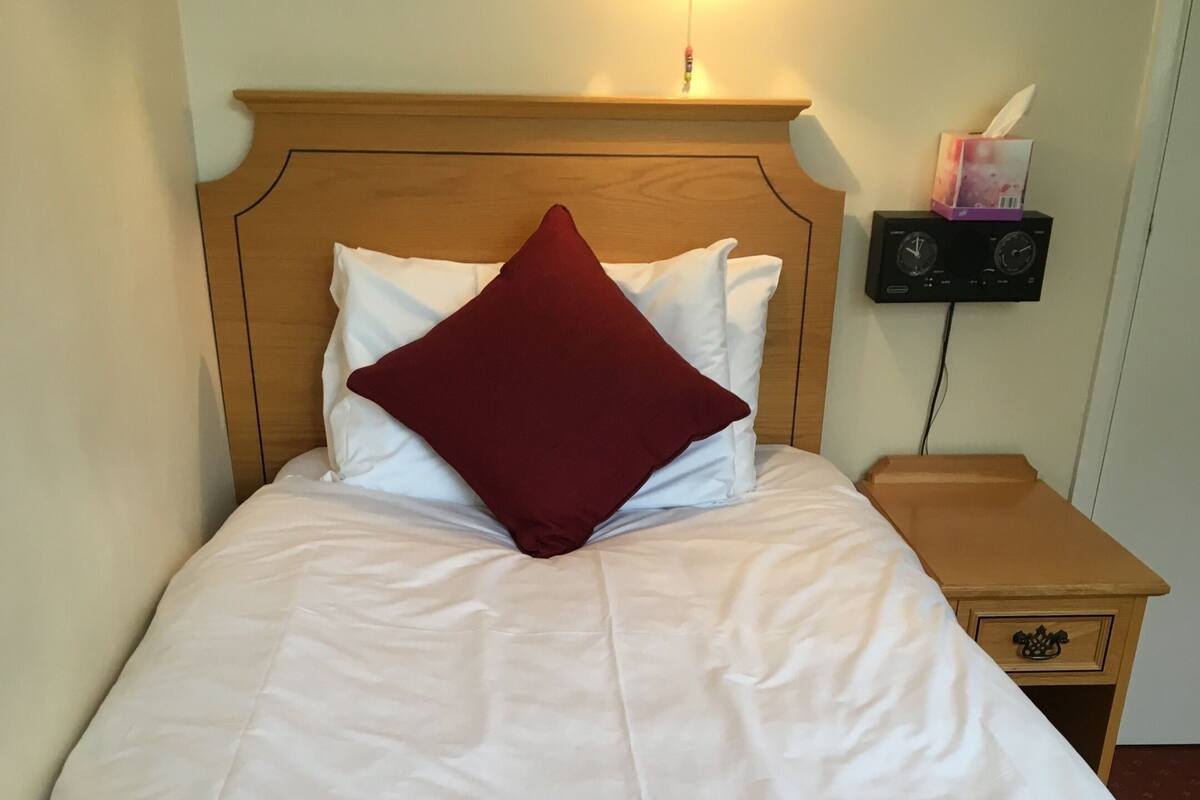 Una cama bien tendida con edredón blanco y manta roja, cabecera de madera y mesita de noche con caja de pañuelos y radio.