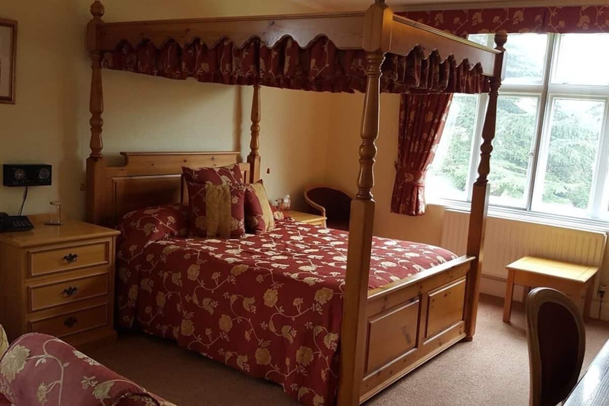 Una cama con dosel y cortinas, un edredón estampado con motivos florales, una mesita de noche de madera, un escritorio y una silla.