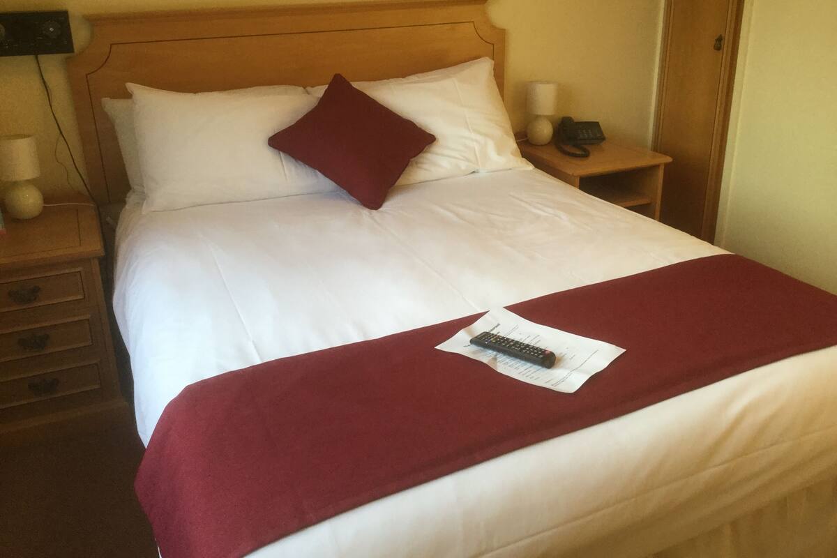 Una cama bien hecha con sábanas blancas y un almohadón color burdeos, un control remoto sobre la cama y una mesita de noche con una lámpara y un teléfono.