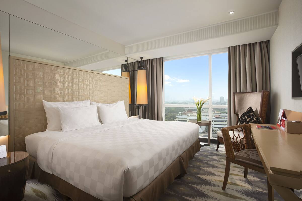 A hotel room with a large bed, a desk, a chair, and a view of the city.