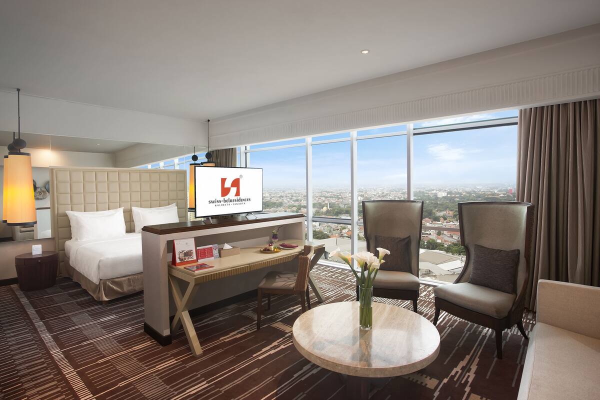 A hotel room with a bed, a desk, two armchairs, a round table, and a large window offering a city view.