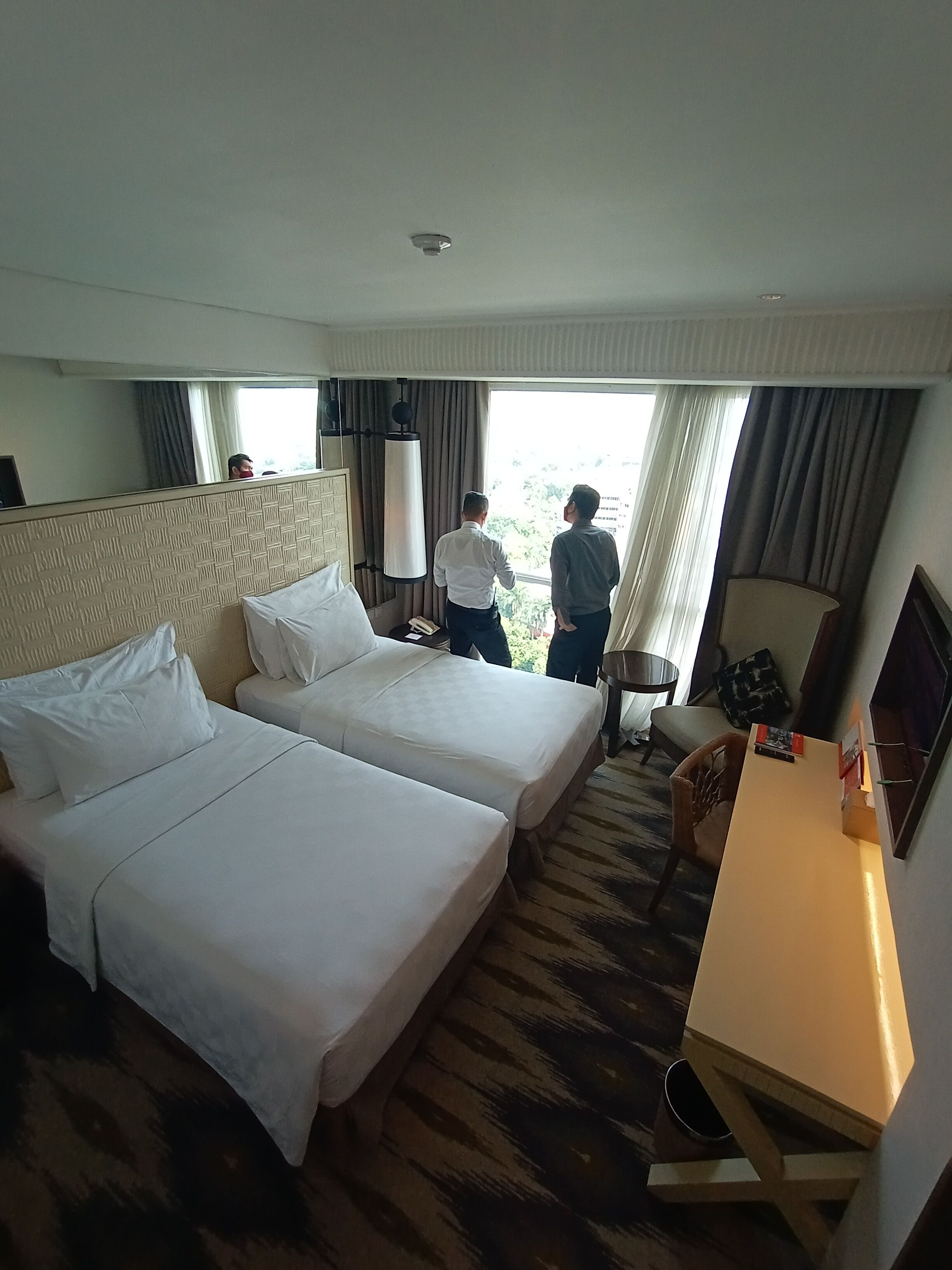 A hotel room with two beds, a desk, a chair, and a large window with curtains.