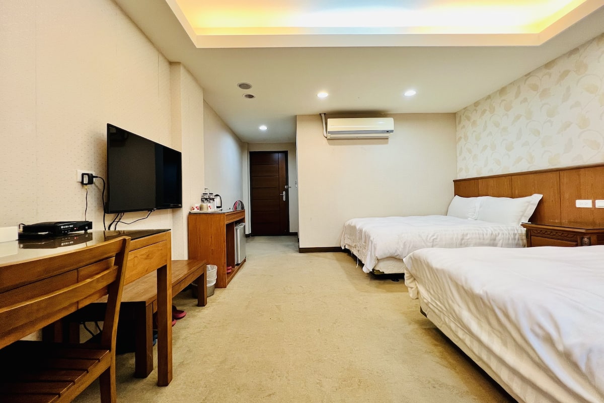 A hotel room with a desk, TV, two beds, and an air conditioning unit.