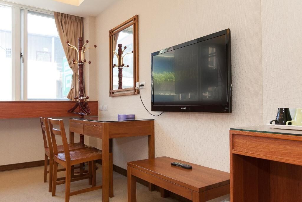 A hotel room with a wooden desk, a television mounted on the wall, a window with curtains, and a small table with a remote control.