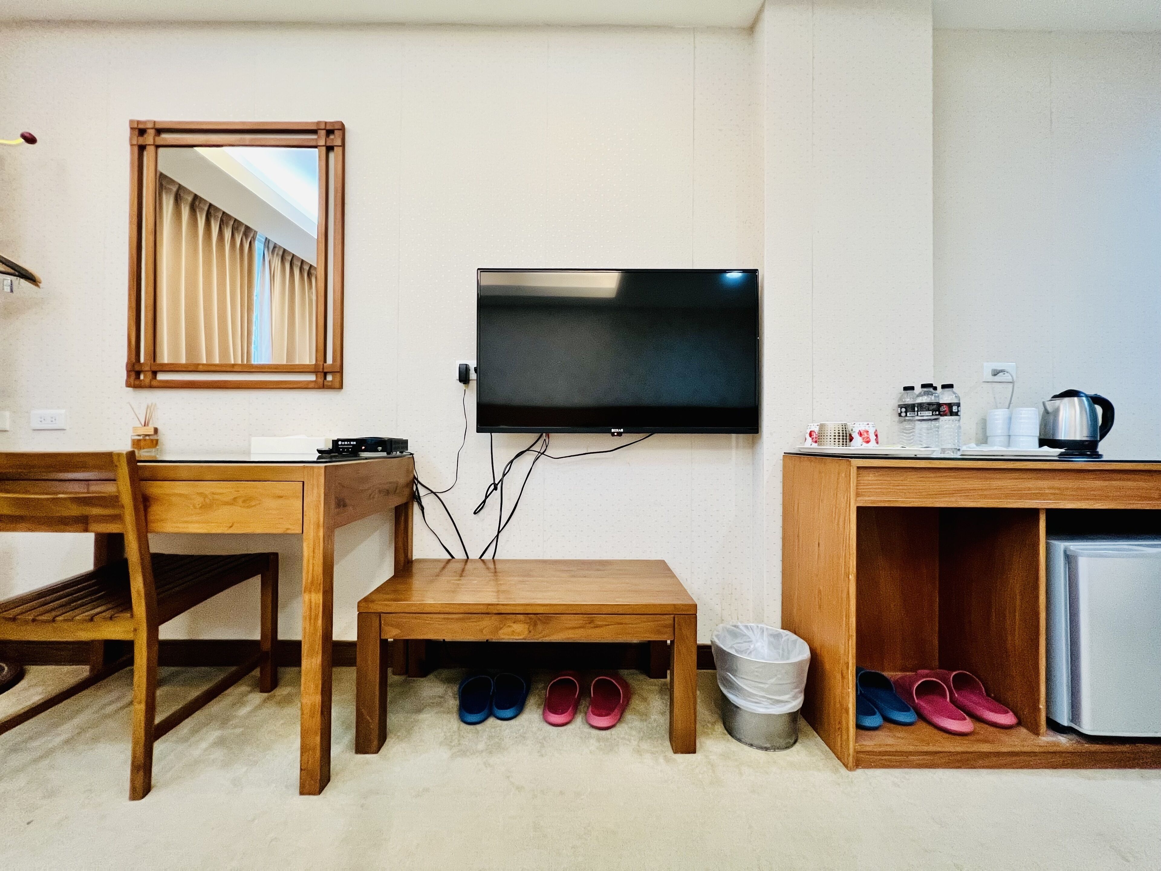 Ein kleines Hotelzimmer mit einem Holzbüro, einem an der Wand befestigten Fernseher, einem kleinen Tisch, einem Spiegel und einem Schuhregal mit Hausschuhen.