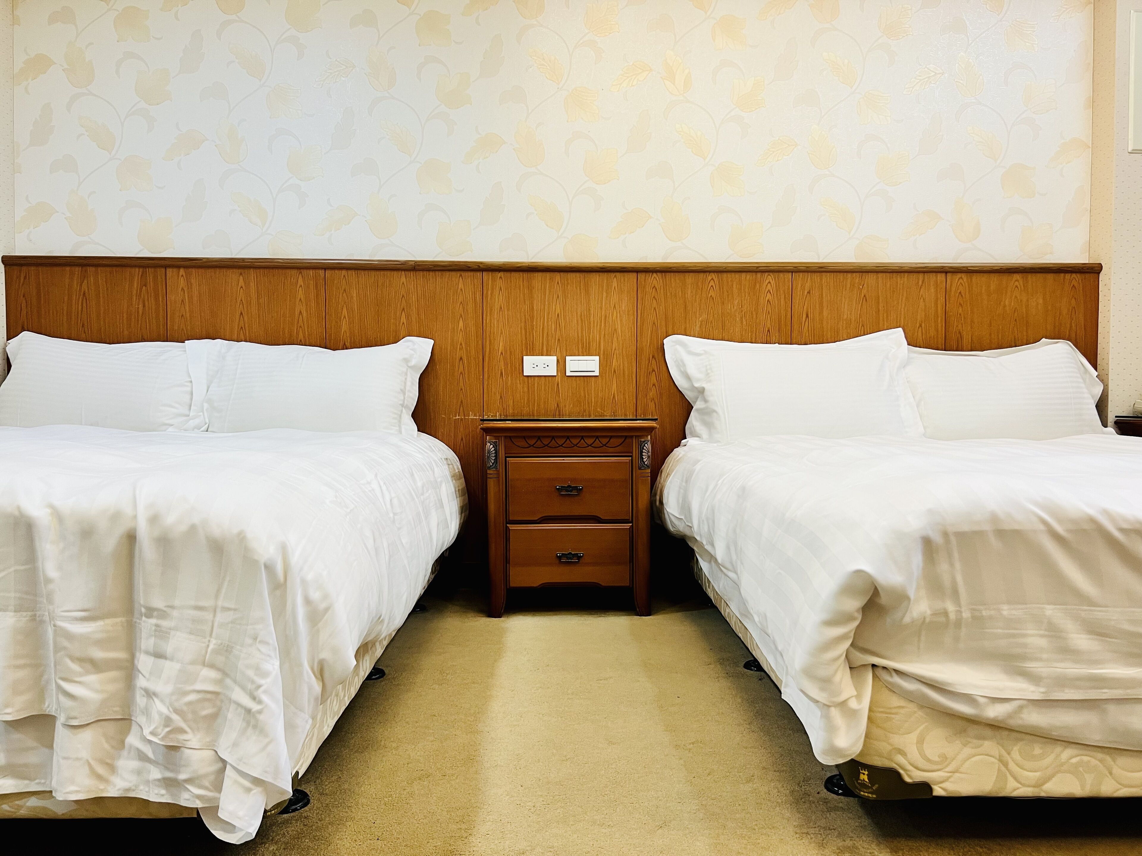 A hotel room with two single beds, a wooden headboard, a nightstand with drawers, and a patterned wallpaper.