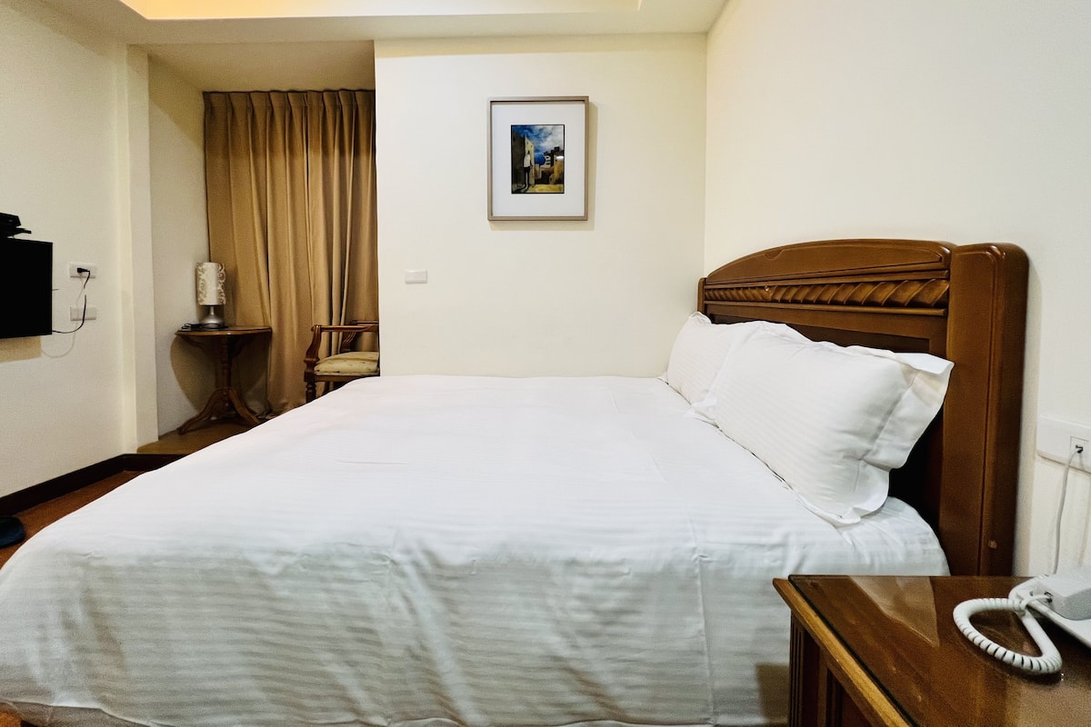 A hotel room with a bed, a bedside table with a lamp, a chair, a framed picture on the wall, and a telephone on the table.