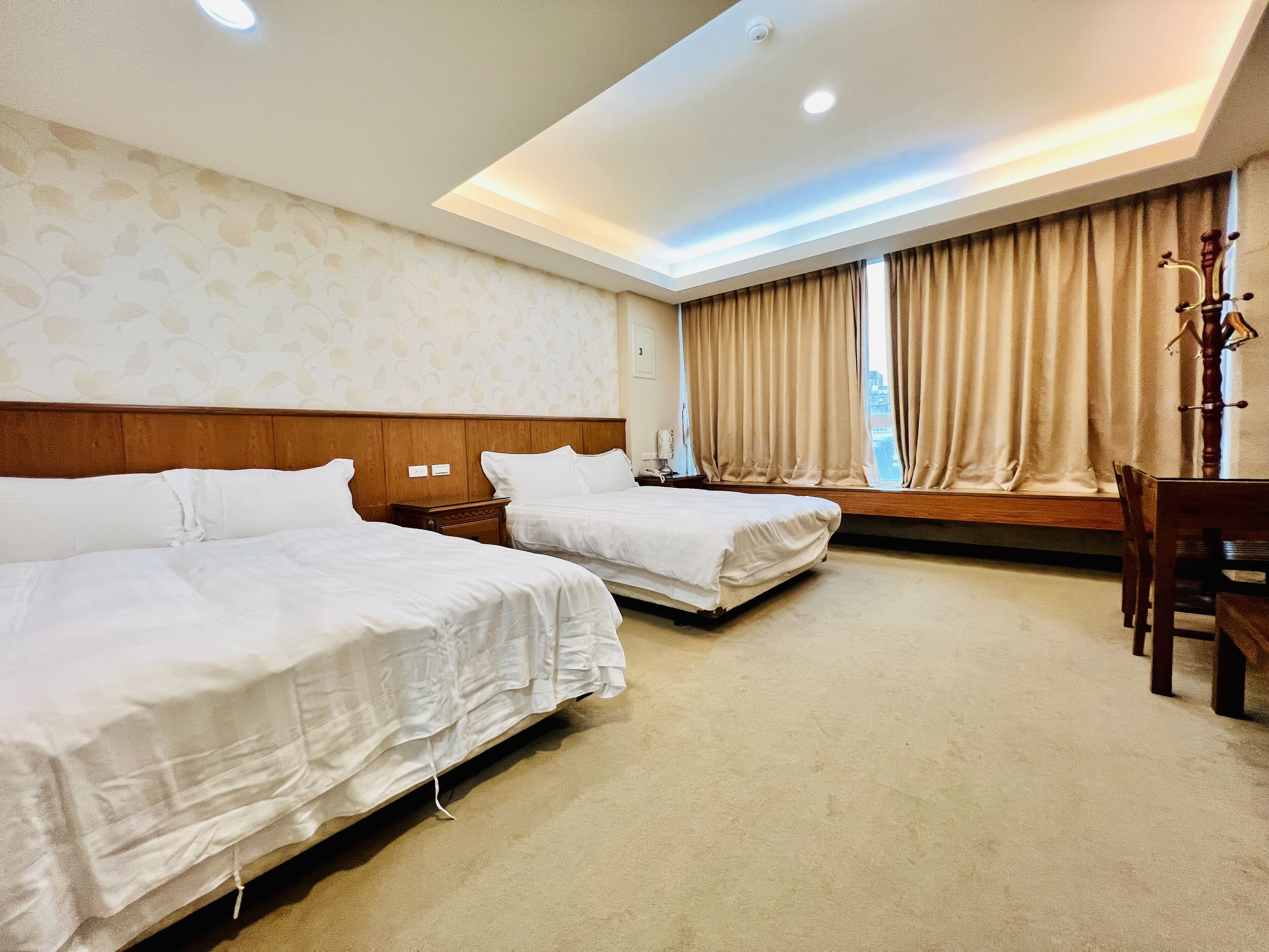 Une chambre d’hôtel avec deux lits, une tête de lit en bois, un bureau avec une chaise et une fenêtre avec des rideaux.