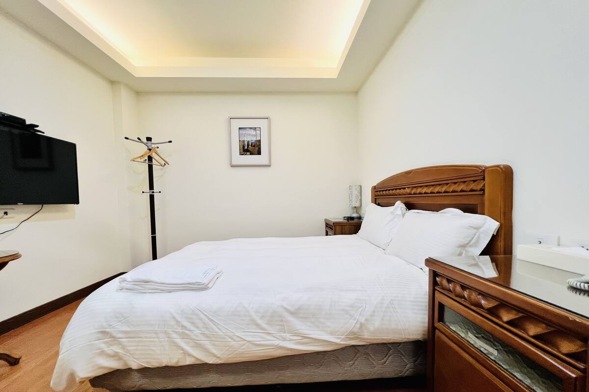 A neatly made bed with white linens, a wooden headboard, a bedside table with a lamp, and a framed picture on the wall.